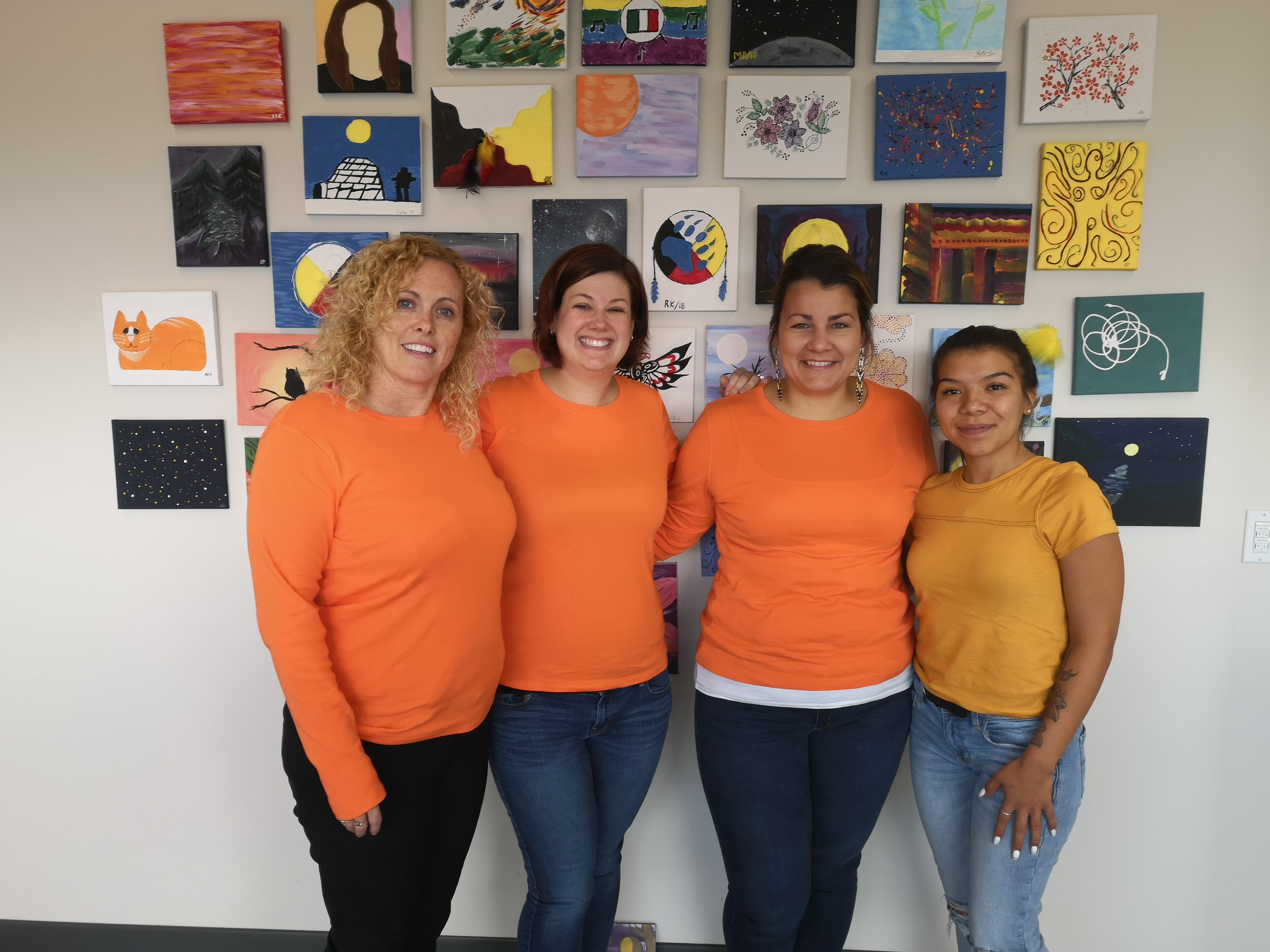 Four women are wearing bright orange shirts and standing in front of a wall of brightly coloured Indigenous art.